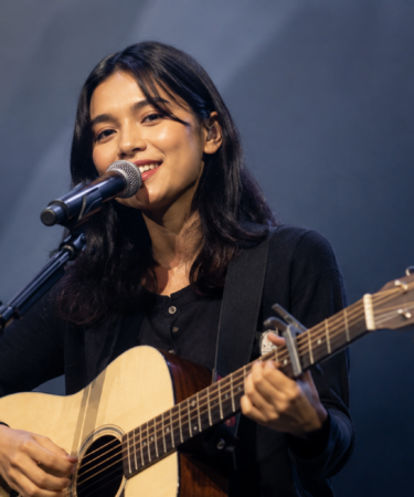 Feby Putri tampil membawakan lagu di atas panggung dengan gitar akustik dan pencahayaan hangat